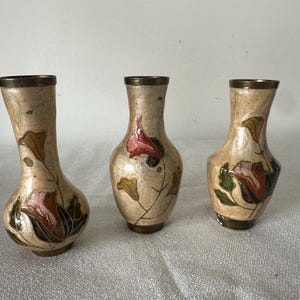 Pode incluir: Três vasos decorativos com fundo bege e bordas e bases de latão. Cada vaso apresenta um desenho floral com detalhes em rosa, verde e castanho. Os vasos variam ligeiramente em forma e tamanho.