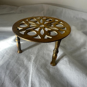 May include: A round gold-toned metal trivet featuring an intricate openwork floral pattern on the top surface. The trivet is supported by three slender, turned metal legs that provide elevation, showcasing a metallic finish throughout the piece.