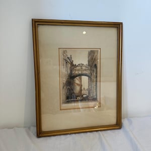 May include: A framed etching depicting the Bridge of Sighs in Venice, Italy. The artwork features detailed architectural lines of the stone bridge spanning a canal, enclosed in a simple gold-toned frame with a cream-colored matting border.