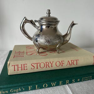 Puede incluir: Una pequeña tetera plateada con detalles florales ornamentados, colocada sobre una pila de libros. La tetera tiene un asa curvada, un pico y una tapa con un pequeño pomo. Los libros se titulan "THE STORY OF ART" y "Van Gogh's FLOWERS".