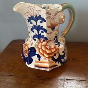 Puede incluir: Jarra de cerámica antigua con un diseño floral en azul, naranja y blanco. La jarra tiene una forma octogonal única y un asa con un diseño verde y naranja. El pico es festoneado.