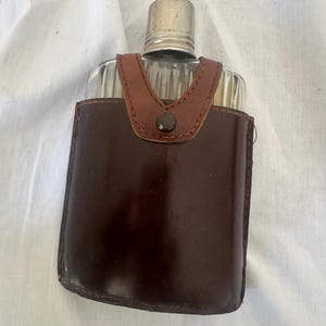 May include: A clear glass flask with vertical ridges, topped with a silver-toned metal screw cap. The flask is encased in a dark brown leather sleeve featuring a V-shaped strap secured by a single dark metal snap button at the centre front.
