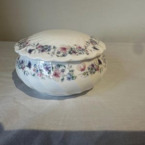 Puede incluir: Una caja de baratijas de cerámica blanca con tapa, adornada con un estampado floral en tonos rosa, morado y azul. La caja tiene un borde festoneado y una forma redondeada, adecuada para guardar objetos pequeños.
