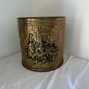 May include: A cylindrical brass bucket featuring an embossed relief scene of figures gathered around a table in a tavern setting. The metal has a polished gold-toned finish with darkened recessed areas that highlight the intricate details of the design.