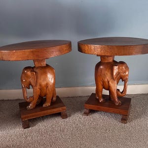 May include: Two wooden side tables with carved elephant bases and round tabletops. The tables are made of brown wood and have a rustic, handcrafted appearance. Each table is approximately 61 cm tall.