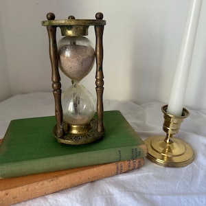 May include: An antique brass hourglass with clear glass bulbs containing light-coloured sand, positioned atop a green hardcover book resting on an orange hardcover book, accompanied by a brass candlestick holder with a white taper candle.