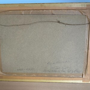 May include: Back of a framed artwork with a gold-colored wooden frame. The artwork is secured with a wire and metal hardware. The back of the artwork is a light brown color. Handwritten text is visible on the back.