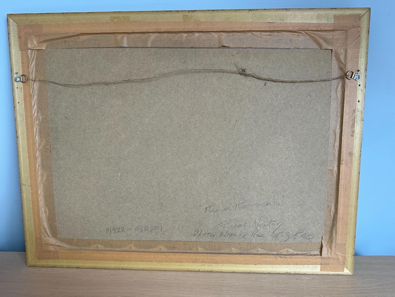 May include: Back of a framed artwork with a gold-colored wooden frame. The artwork is secured with a wire and metal hardware. The back of the artwork is a light brown color. Handwritten text is visible on the back.