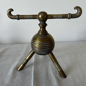 May include: A vintage brass stand with a tripod base and a horizontal bar with curved ends. The stand has a spherical center section with horizontal grooves and decorative detailing. The brass has a warm, aged patina.
