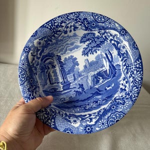 May include: A blue and white decorative plate with a landscape scene. The plate features a detailed illustration of a classical landscape with figures, trees, and buildings. The rim of the plate has a floral pattern.