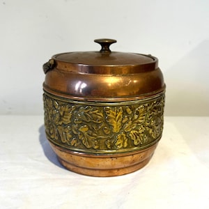 May include: A copper and brass container with a lid. The container has a cylindrical shape with a decorative band of embossed leaves and vines. The lid has a small knob on top. The container is likely vintage and has a rustic appearance.