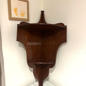 May include: A dark brown wooden corner shelf with two shelves and a decorative top, mounted on a white wall. A framed picture with handprints and the name "Wilfred" is visible.