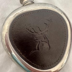 Op de afbeelding: Een zilverkleurige metalen heupfles met een ronde vorm en een getextureerde schroefdop. Het midden is voorzien van een donkerbruine, schildvormige lederen inleg met een reliëf van een hertenkop met groot, vertakt gewei.