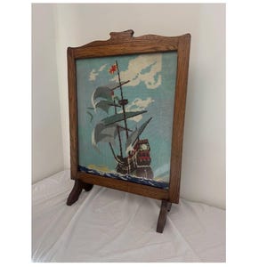 May include: A rectangular wooden fire screen featuring a needlepoint tapestry of a sailing ship on a light blue background with white clouds. The ship has dark sails and a red flag, set against a sea with dark blue waves and a yellow shoreline.