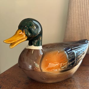 May include: A ceramic duck figurine with a green head, brown body, and orange beak. The duck is sitting on a wooden surface.