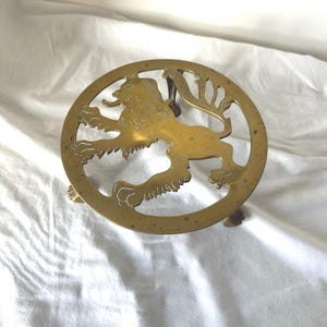 Può includere: Un sottopentola circolare in ottone con un design traforato raffigurante un leone rampante. L'oggetto in metallo presenta una finitura dorata lucida con patina visibile e poggia su tre piccoli piedini, con dettagli incisi sulla figura.