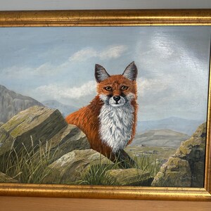 May include: Oil painting of a red fox with a white chest, framed in a gold-colored ornate frame. The fox is positioned between large rocks, with a mountainous landscape and cloudy sky in the background. The artwork is signed.