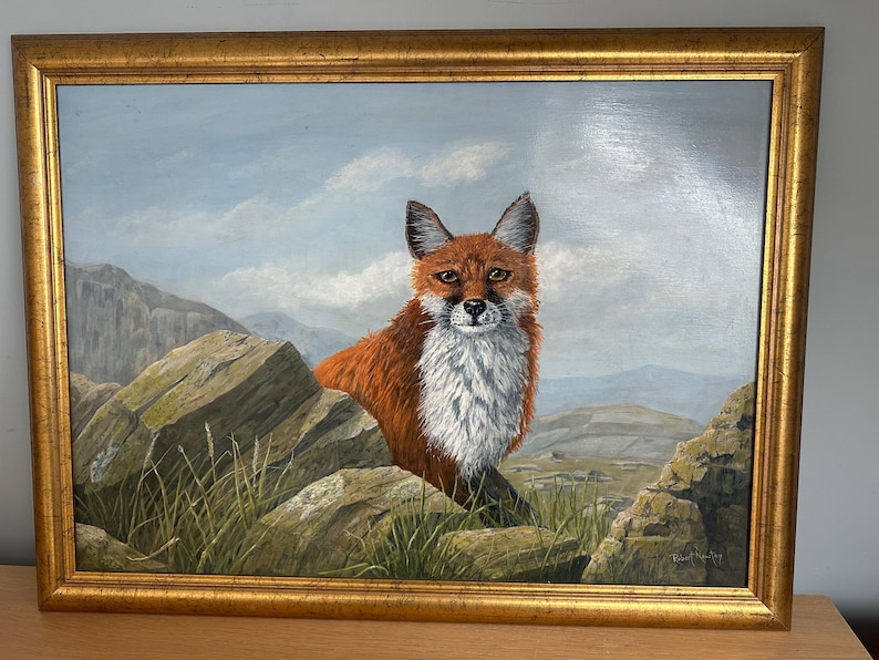 May include: Oil painting of a red fox with a white chest, framed in a gold-colored ornate frame. The fox is positioned between large rocks, with a mountainous landscape and cloudy sky in the background. The artwork is signed.