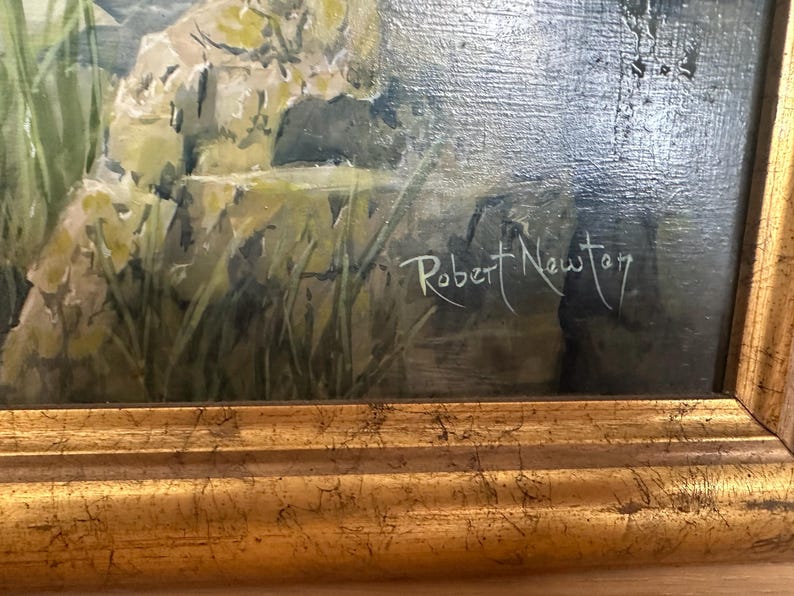 May include: An oil painting depicting a landscape scene with a body of water, rocks, and greenery. The artwork is signed "Robert Newton" in white script. The painting is framed in a gold-colored, ornate frame, adding a touch of elegance.