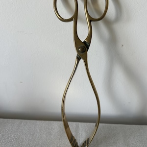 Puede incluir: Pinzas de azúcar vintage de latón. Presentan bucles ovalados para los dedos y un diseño en forma de tijera. El cuerpo curvado conduce a un extremo de agarre estriado. El latón tiene un tono dorado cálido.