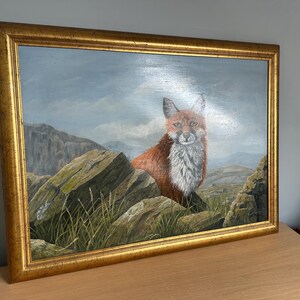 May include: Oil painting of a fox in a gold frame. The fox has red fur and white chest fur, perched on rocks with green grass. The background features a mountain range and a cloudy sky.