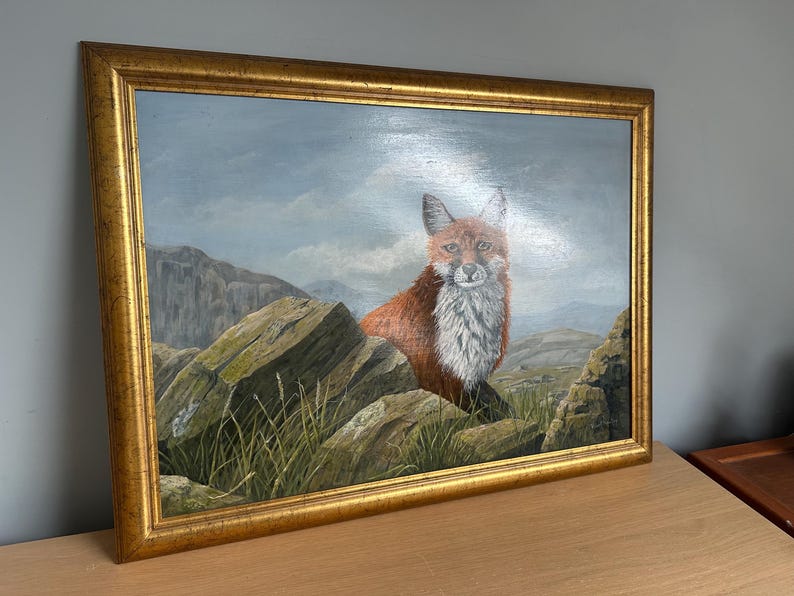 May include: Oil painting of a fox in a gold frame. The fox has red fur and white chest fur, perched on rocks with green grass. The background features a mountain range and a cloudy sky.