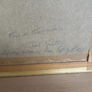 May include: A framed artwork with the title "Fox on the rocks" handwritten in black ink. The artist's name, Robert Newton, and address are also visible. The frame is gold-colored.