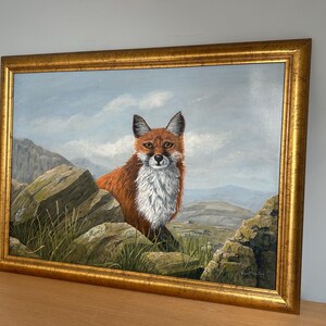 May include: A framed oil painting of a fox in a mountainous landscape. The fox has reddish-brown fur and a white chest, set against a backdrop of rocks, grass, and a cloudy sky. The frame is gold-colored.