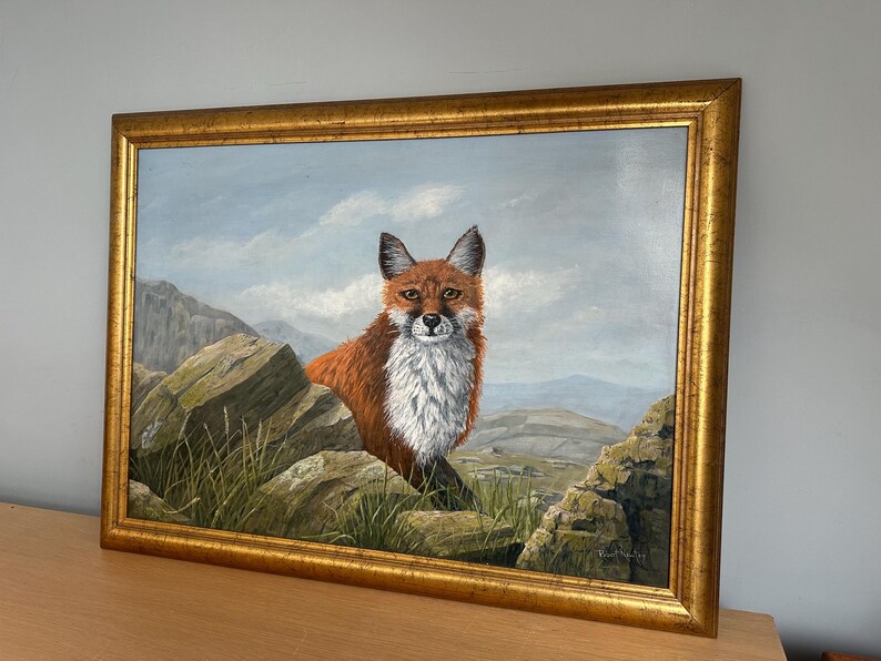 May include: A framed oil painting of a fox in a mountainous landscape. The fox has reddish-brown fur and a white chest, set against a backdrop of rocks, grass, and a cloudy sky. The frame is gold-colored.