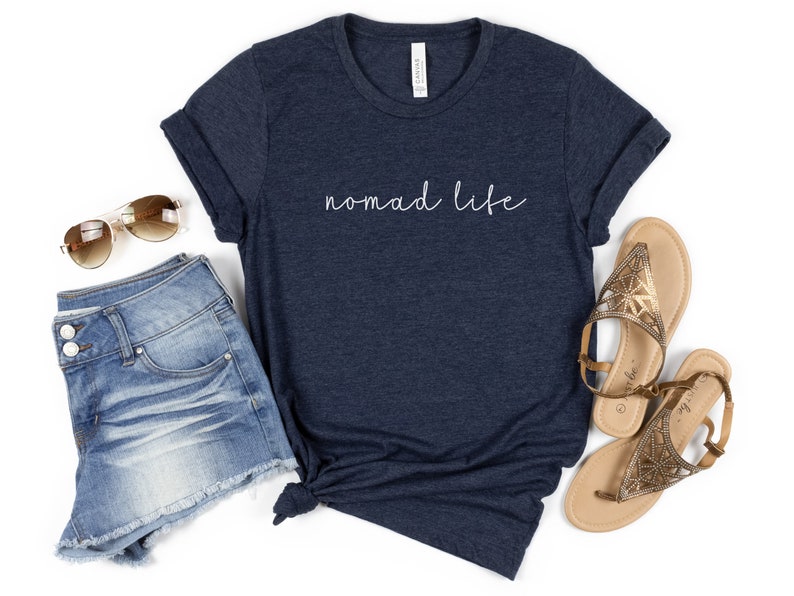 K&ouml;nnte beinhalten: Ein marineblaues T-Shirt mit dem Text "Nomad Life" in wei&szlig;er Schrift. Das Shirt wird mit Jeans-Shorts und goldenen Sandalen kombiniert.