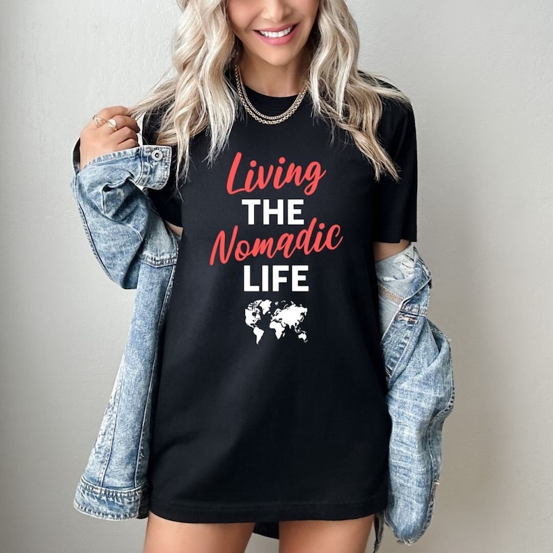 Op de afbeelding: Zwart T-shirt met de tekst "Living the Nomadic Life" in rode letters en een witte omtrek van een wereldkaart.