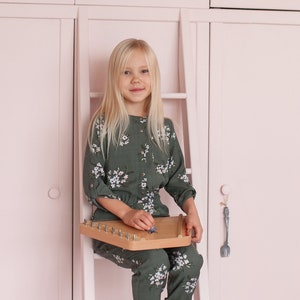 May include: A young girl sits on a white wooden ladder in front of a pink cabinet. She is wearing a green floral jumpsuit and white socks. She is holding a wooden musical instrument.
