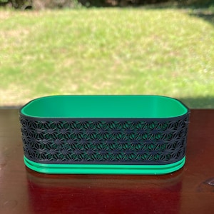 Könnte beinhalten: Ein rechteckiger Behälter in Grün und Schwarz mit einem geometrischen Muster. Der Behälter ist aus Kunststoff und hat eine glatte Oberfläche.