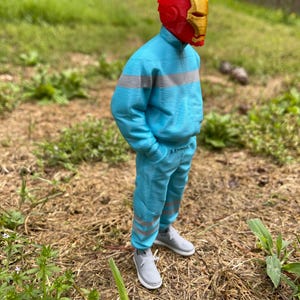 May include: A figure wearing a red and gold Iron Man helmet, a light blue tracksuit with gray stripes, and gray sneakers. The figure is standing outdoors on a bed of dry grass and weeds, with green grass and a fence in the background.