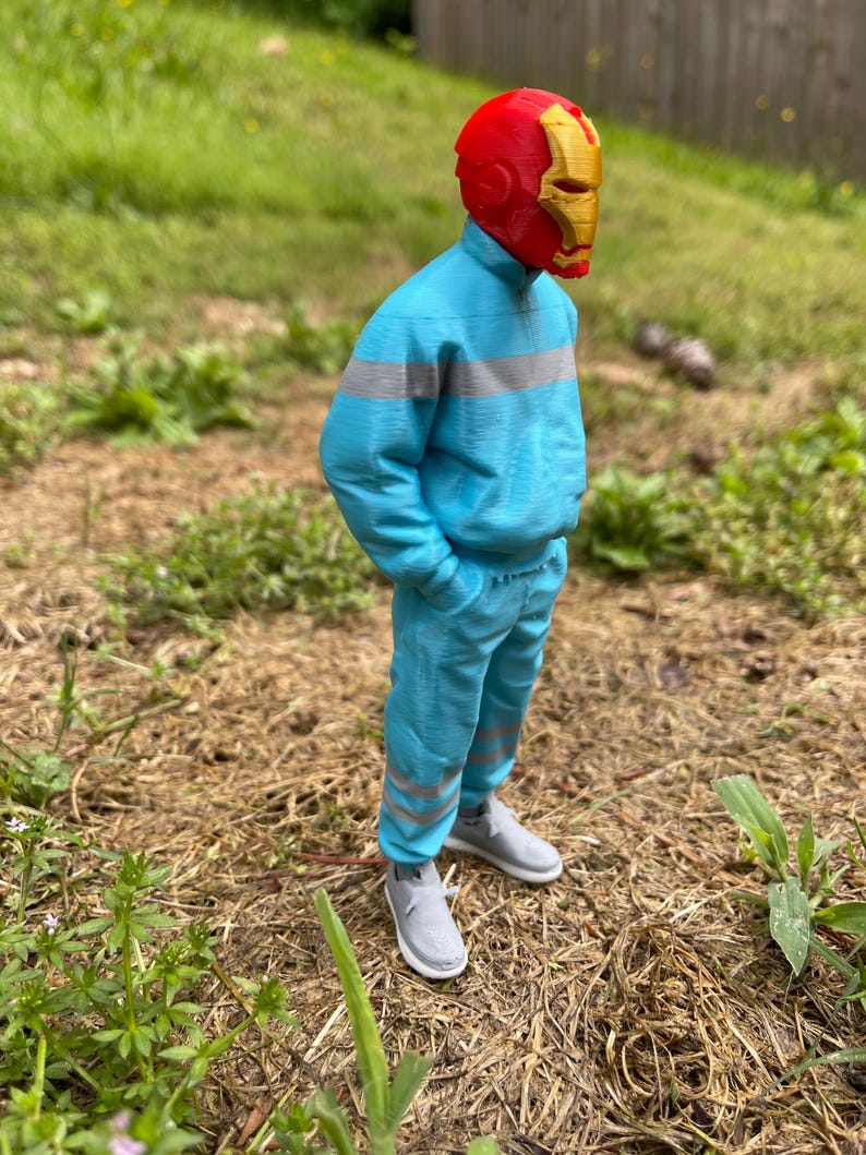May include: A figure wearing a red and gold Iron Man helmet, a light blue tracksuit with gray stripes, and gray sneakers. The figure is standing outdoors on a bed of dry grass and weeds, with green grass and a fence in the background.