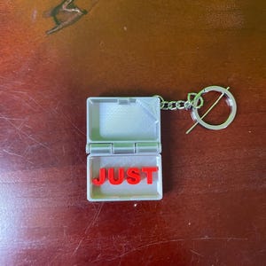 Op de afbeelding: Een kleine, grijze, scharnierende sleutelhanger met het woord "JUST" in rode letters erin. De sleutelhanger heeft een zilverkleurige ring en ketting bevestigd aan de doos.