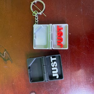 Op de afbeelding: Een zilverkleurige sleutelhanger met een kleine, scharnierende doos. E&eacute;n doos is open en toont het woord "JUST" in rode letters. De andere doos is gesloten, met "JUST" in witte letters. De sleutelhanger ligt op een bruin oppervlak.