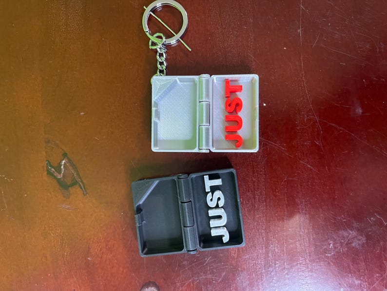 Op de afbeelding: Een zilverkleurige sleutelhanger met een kleine, scharnierende doos. E&eacute;n doos is open en toont het woord "JUST" in rode letters. De andere doos is gesloten, met "JUST" in witte letters. De sleutelhanger ligt op een bruin oppervlak.