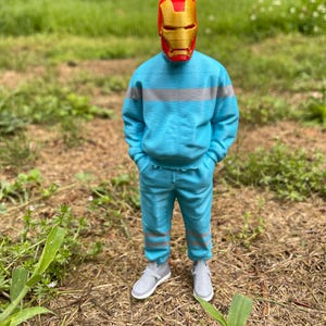 May include: A person wearing a red and gold Iron Man mask, a light blue sweatshirt with gray stripes, matching pants, and gray sneakers. The person is standing in a grassy area with brown mulch and green plants.