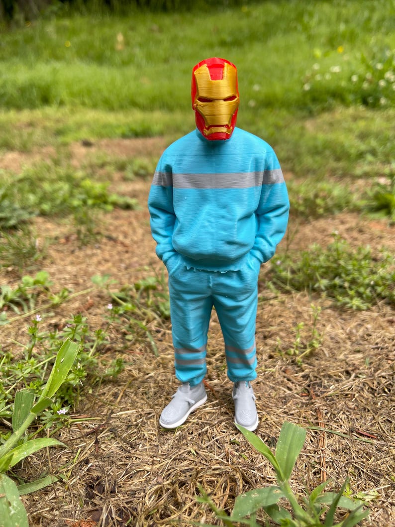 May include: A person wearing a red and gold Iron Man mask, a light blue sweatshirt with gray stripes, matching pants, and gray sneakers. The person is standing in a grassy area with brown mulch and green plants.