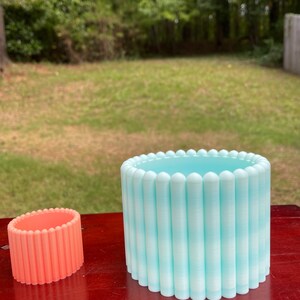 Könnte beinhalten: Zwei Keramik-Blumentöpfe, einer hellblau und einer korallenfarben, mit einem Gingham-Muster. Die Töpfe sind zylindrisch und haben einen erhöhten Rand.