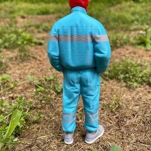 May include: A 3D-printed figure of a person wearing a red hat, a light blue jacket with gray stripes, and matching pants. The figure is wearing gray sneakers. The background is a grassy area.