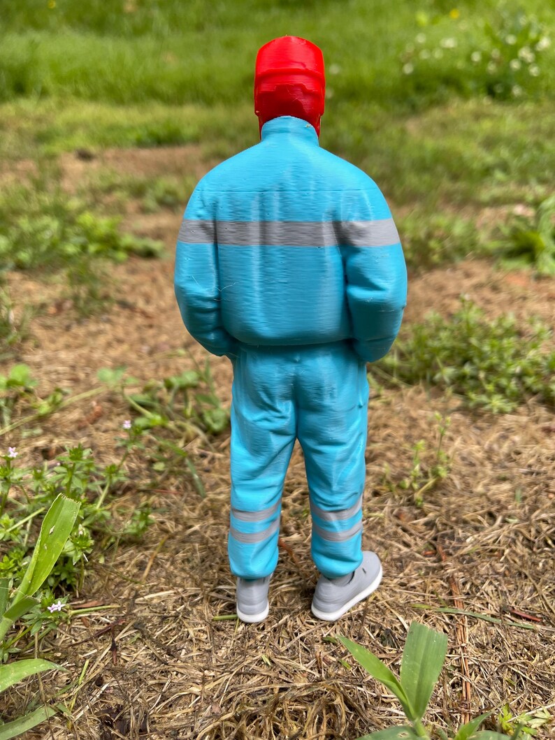 May include: A 3D-printed figure of a person wearing a red hat, a light blue jacket with gray stripes, and matching pants. The figure is wearing gray sneakers. The background is a grassy area.