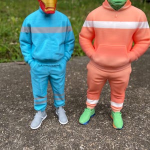 May include: Two figures dressed in superhero-themed outfits. One figure wears a red and gold Iron Man mask with a blue sweatsuit. The other figure is a green Hulk head with an orange sweatsuit. Both figures wear sneakers.