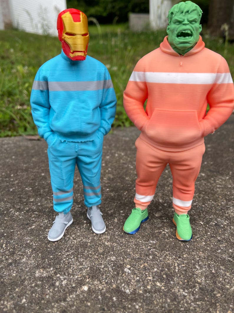 May include: Two figures dressed in superhero-themed outfits. One figure wears a red and gold Iron Man mask with a blue sweatsuit. The other figure is a green Hulk head with an orange sweatsuit. Both figures wear sneakers.