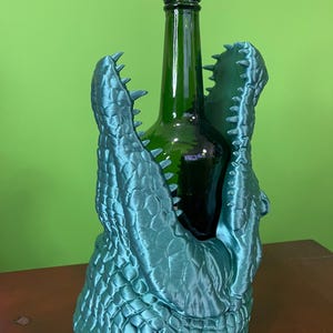 Op de afbeelding: Een teal-kleurige wijnfleshouder in de vorm van een alligator. De houder is ontworpen om een groene wijnfles te omarmen, met de kaken van de alligator open. De houder heeft een getextureerd, geschubd uiterlijk.