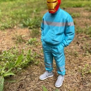 May include: A figure wearing a red and gold Iron Man mask, a light blue tracksuit with gray stripes, and gray sneakers stands outdoors. The figure is in a grassy area with brown ground cover.