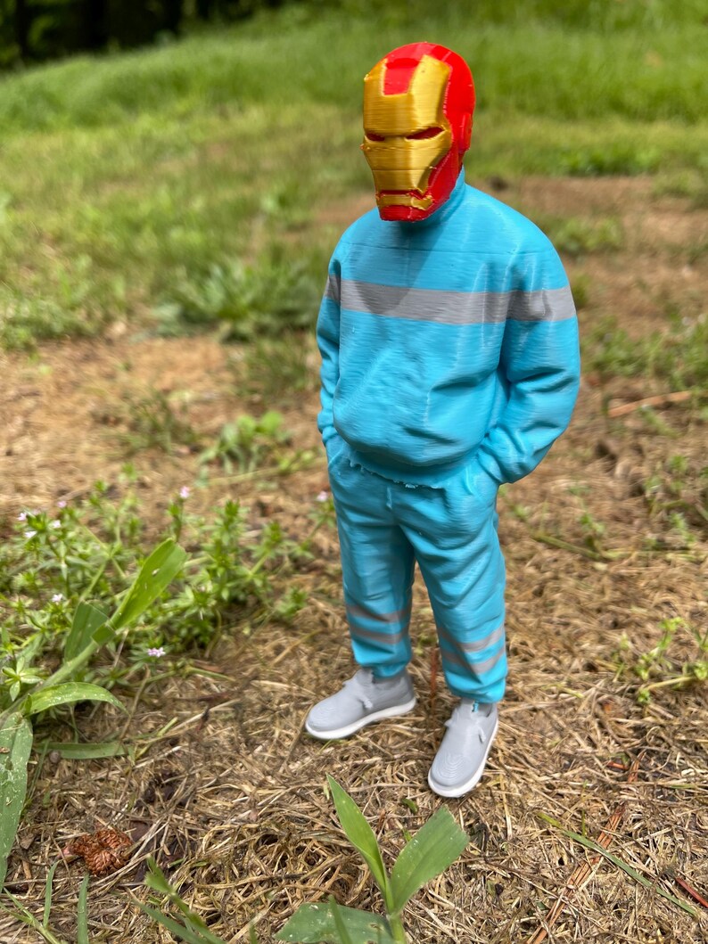 May include: A figure wearing a red and gold Iron Man mask, a light blue tracksuit with gray stripes, and gray sneakers stands outdoors. The figure is in a grassy area with brown ground cover.