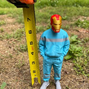 May include: A 3D-printed figure of a superhero wearing a red and gold mask, blue sweatsuit, and gray sneakers. The figure is approximately 5 inches tall. The figure is standing in a grassy area, with a measuring tape held next to it.