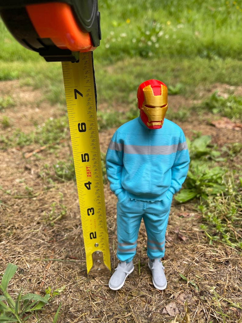 May include: A 3D-printed figure of a superhero wearing a red and gold mask, blue sweatsuit, and gray sneakers. The figure is approximately 5 inches tall. The figure is standing in a grassy area, with a measuring tape held next to it.